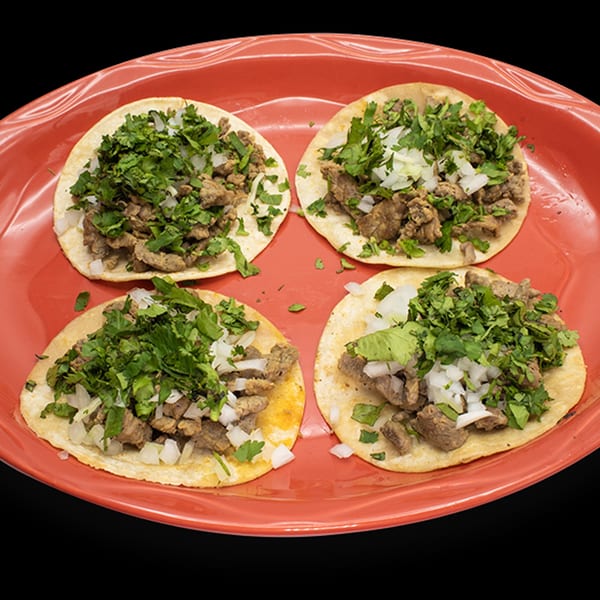 Four authentic Mexican street tacos with carne asada, cilantro, and diced onions on a red plate.